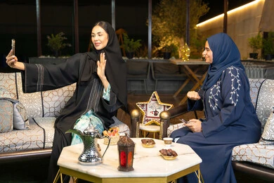 حسن الاستقبال وكرم الضيافة في شهر رمضان المبارك، امراتان سعوديتان تقومان بتوثيق اللحظات السعيدة، التقاط صور سيلفي باستخدام الهاتف المحمول، صديقتان عربيتان خليجينان اماراتيتان ترتديان العباءة تجلسان في الهواء الطلق، زينة واضاءة رمضانية، الجلسة الليلة الخارجية