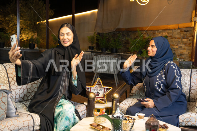امراتان سعوديتان تقومان بتوثيق اللحظات السعيدة، التقاط صور سيلفي باستخدام الهاتف المحمول، حسن الاستقبال وكرم الضيافة في شهر رمضان المبارك، صديقتان عربيتان خليجينان اماراتيتان ترتديان العباءة تجلسان في الهواء الطلق، زينة واضاءة رمضانية، الجلسة الليلة الخارجية