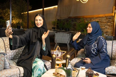 Two Saudi women documenting happy moments, taking selfies with their mobile phones, the warm welcome and hospitality during the holy month of Ramadan, two Emirati Gulf Arab friends wearing abayas sitting outdoors, Ramadan decorations and lighting, outdoor night session
