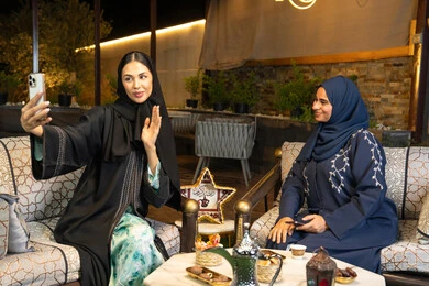 التقاط صور سيلفي باستخدام الهاتف المحمول، امراتان سعوديتان تقومان بتوثيق اللحظات السعيدة، حسن الاستقبال وكرم الضيافة في شهر رمضان المبارك، صديقتان عربيتان خليجينان اماراتيتان ترتديان العباءة تجلسان في الهواء الطلق، زينة واضاءة رمضانية، الجلسة الليلة الخارجية