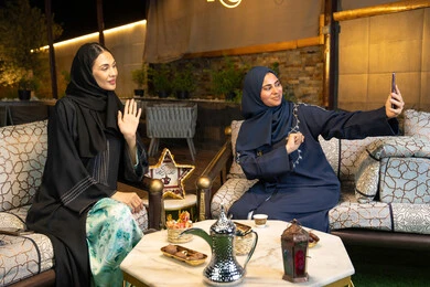 امراتان سعوديتان تقومان بتوثيق اللحظات السعيدة، التقاط صور سيلفي باستخدام الهاتف المحمول، حسن الاستقبال وكرم الضيافة في شهر رمضان المبارك، صديقتان عربيتان خليجينان اماراتيتان ترتديان العباءة تجلسان في الهواء الطلق، زينة واضاءة رمضانية، الجلسة الليلة الخارجية
