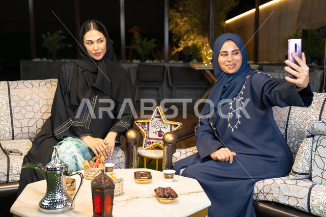 كرم الضيافة وحسن الاستقبال في شهر رمضان المبارك، امراتان سعوديتان تقومان بتوثيق اللحظات السعيدة، التقاط صور سيلفي باستخدام الهاتف المحمول، صديقتان عربيتان خليجينان اماراتيتان ترتديان العباءة تجلسان في الهواء الطلق، زينة واضاءة رمضانية، الجلسة الليلة الخارجية