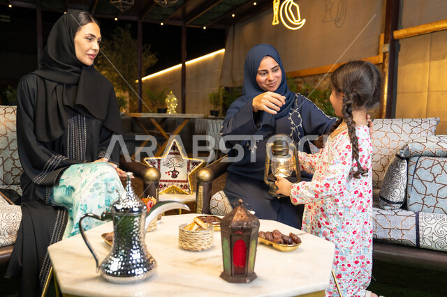 حسن الاستقبال وكرم الضيافة في شهر رمضان المبارك، زينة واضاءة رمضانية، الجلسة الليلة الخارجية في احدى شاليهات الامارات،  امراة عربية خليجية اماراتية تجلس في الهواء الطلق مع صديقتها وابنتها، سيدتان سعوديتان تقومان بتبادل الاحاديث والاخبار بتعابير السرور