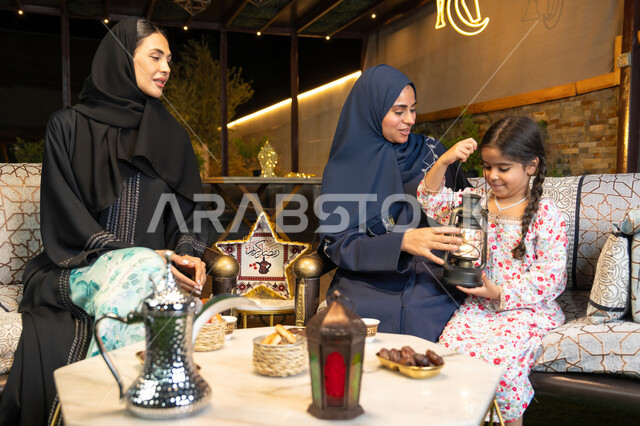 جودة الاستقبال وكرم الضيافة في شهر رمضان المبارك، زينة واضاءة رمضانية، الجلسة الليلة الخارجية في احدى شاليهات الامارات،  امراة عربية خليجية اماراتية تجلس في الهواء الطلق مع صديقتها وابنتها، سيدتان سعوديتان تقومان بتبادل الاحاديث والاخبار بتعابير السرور