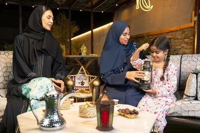 جودة الاستقبال وكرم الضيافة في شهر رمضان المبارك، زينة واضاءة رمضانية، الجلسة الليلة الخارجية في احدى شاليهات الامارات،  امراة عربية خليجية اماراتية تجلس في الهواء الطلق مع صديقتها وابنتها، سيدتان سعوديتان تقومان بتبادل الاحاديث والاخبار بتعابير السرور