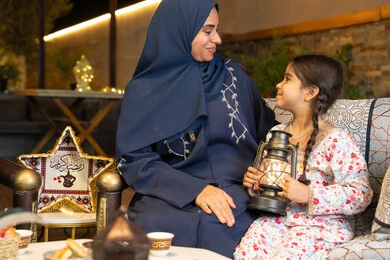 An outdoor evening session in one of the chalets in the Emirates, celebrating the arrival of the month of goodness, decorations and lighting for the holy month of Ramadan, an Emirati Gulf Arab woman sitting outdoors, a Saudi woman looking at her daughter with gestures of happiness and pleasure