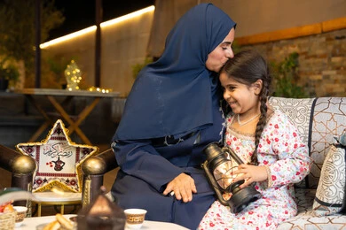 Decorations and lighting for the holy month of Ramadan, an outdoor evening session in one of the chalets in the Emirates, an Emirati Gulf Arab woman sitting outdoors, a Saudi woman kissing her daughter, a girl holding a Ramadan lantern in her hand