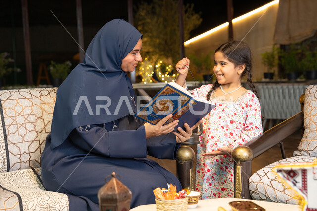 تعزيز الفطرة الاسلامية لدى الاولاد، تلاوة وقراءة كتاب الله تعالى ,الإجتهاد و الخشوع في العباده, ام عربية سعودية خليجية ترتدي الحجاب و العباءة تمسك بيدها القرآن الكريم برفقة ابنتها، سيدة اماراتية تجلس على الكنبة في الهواء الطلق, التقرب الى الله في شهر رمضان المبارك