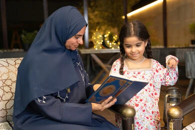 تقوية الفطرة الاسلامية لدى الاولاد، تلاوة وقراءة كتاب الله تعالى ,الإجتهاد و الخشوع في العباده, ام عربية سعودية خليجية ترتدي الحجاب و العباءة تمسك بيدها القرآن الكريم برفقة ابنتها، سيدة اماراتية تجلس على الكنبة في الهواء الطلق, التقرب الى الله في شهر رمضان المبارك