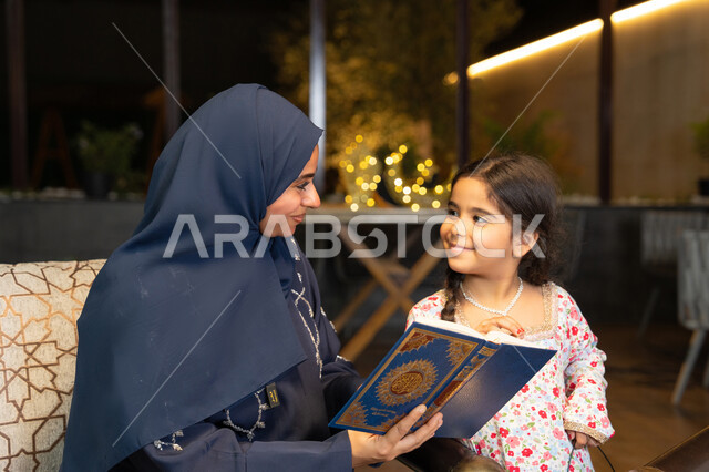 تعزيز الفطرة الاسلامية لدى الاولاد، تلاوة وقراءة كتاب الله تعالى ,الإجتهاد و الخشوع في العباده, ام عربية سعودية خليجية ترتدي الحجاب و العباءة تمسك بيدها القرآن الكريم برفقة ابنتها، سيدة اماراتية تجلس على الكنبة في الهواء الطلق, التقرب الى الله في شهر رمضان المبارك