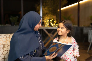 تعزيز الفطرة الاسلامية لدى الاولاد، تلاوة وقراءة كتاب الله تعالى ,الإجتهاد و الخشوع في العباده, ام عربية سعودية خليجية ترتدي الحجاب و العباءة تمسك بيدها القرآن الكريم برفقة ابنتها، سيدة اماراتية تجلس على الكنبة في الهواء الطلق, التقرب الى الله في شهر رمضان المبارك