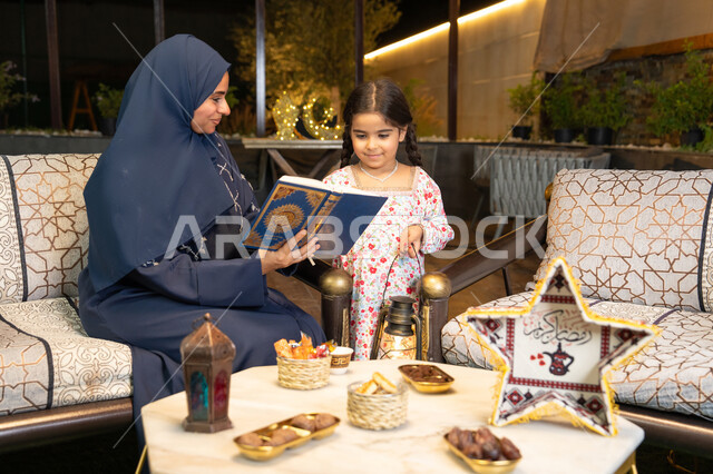 تلاوة وقراءة كتاب الله تعالى ,الإجتهاد و الخشوع في العباده, ام عربية سعودية خليجية ترتدي الحجاب و العباءة تمسك بيدها القرآن الكريم برفقة ابنتها، سيدة اماراتية تجلس على الكنبة في الهواء الطلق, التقرب الى الله في شهر رمضان المبارك، تعزيز الفطرة الاسلامية لدى الاولاد
