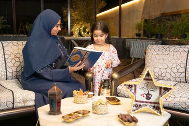 تلاوة وقراءة كتاب الله تعالى ,الإجتهاد و الخشوع في العباده, ام عربية سعودية خليجية ترتدي الحجاب و العباءة تمسك بيدها القرآن الكريم برفقة ابنتها، سيدة اماراتية تجلس على الكنبة في الهواء الطلق, التقرب الى الله في شهر رمضان المبارك، تعزيز الفطرة الاسلامية لدى الاولاد