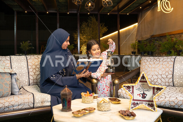 Reciting and reading the Book of God Almighty, diligence and humility in worship, a Saudi Arabian Gulf mother wearing the hijab and abaya holding the Holy Quran in her hand with her daughter, an Emirati woman sitting on a couch in the open air, getting closer to God in the holy month of Ramadan, strengthening the Islamic nature in children