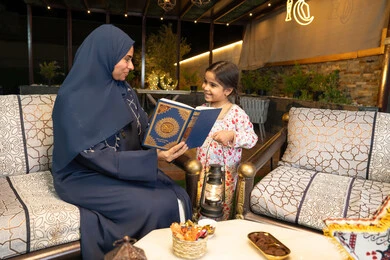 تعزيز الفطرة الاسلامية لدى الاولاد، تلاوة وقراءة كتاب الله تعالى ,الإجتهاد و الخشوع في العباده, ام عربية سعودية خليجية ترتدي الحجاب و العباءة تمسك بيدها القرآن الكريم برفقة ابنتها، سيدة اماراتية تجلس على الكنبة في الهواء الطلق, التقرب الى الله في شهر رمضان المبارك