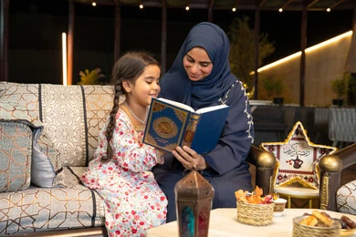 تلاوة وقراءة كتاب الله تعالى ,الإجتهاد و الخشوع في العباده, ام عربية سعودية خليجية ترتدي الحجاب و العباءة تمسك بيدها القرآن الكريم برفقة ابنتها، سيدة اماراتية تجلس على الكنبة في الهواء الطلق, التقرب الى الله في شهر رمضان المبارك، تعزيز الفطرة الاسلامية لدى الاولاد