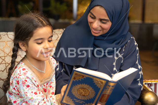 تقوية الفطرة الاسلامية لدى الاولاد، تلاوة وقراءة كتاب الله تعالى ,الإجتهاد و الخشوع في العباده, ام عربية سعودية خليجية ترتدي الحجاب و العباءة تمسك بيدها القرآن الكريم برفقة ابنتها، سيدة اماراتية تجلس على الكنبة في الهواء الطلق, التقرب الى الله في شهر رمضان المبارك