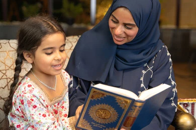 تقوية الفطرة الاسلامية لدى الاولاد، تلاوة وقراءة كتاب الله تعالى ,الإجتهاد و الخشوع في العباده, ام عربية سعودية خليجية ترتدي الحجاب و العباءة تمسك بيدها القرآن الكريم برفقة ابنتها، سيدة اماراتية تجلس على الكنبة في الهواء الطلق, التقرب الى الله في شهر رمضان المبارك