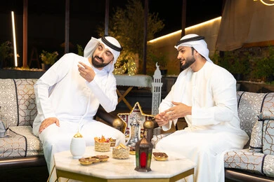 تبادل الاحاديث والاخبار بتعابير السرور، زينة واضاءة رمضانية، الجلسة الليلة الخارجية في احدى شاليهات الامارات، حسن الاستقبال والكرم في شهر رمضان المبارك، صديقان عربيان خليجيان اماراتيان يرتديان الكندورة والغترة يجلسان في الهواء الطلق