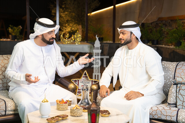 Exchanging conversations and news with expressions of joy, Ramadan decorations and lighting, an outdoor evening gathering in one of the chalets in the Emirates, warm welcome and generosity in the holy month of Ramadan, two Emirati Gulf Arab friends wearing kanduras and ghutras sitting outdoors