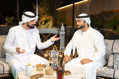 مبادلة الاحاديث والاخبار بتعابير السرور، زينة واضاءة رمضانية، الجلسة الليلة الخارجية في احدى شاليهات الامارات، حسن الاستقبال والكرم في شهر رمضان المبارك، صديقان عربيان خليجيان اماراتيان يرتديان الكندورة والغترة يجلسان في الهواء الطلق