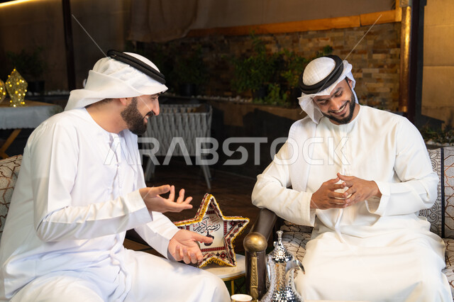 تبادل الاحاديث والاخبار بتعابير السرور، زينة واضاءة رمضانية، الجلسة الليلة الخارجية في احدى شاليهات الامارات، حسن الاستقبال والكرم في شهر رمضان المبارك، صديقان عربيان خليجيان اماراتيان يرتديان الكندورة والغترة يجلسان في الهواء الطلق