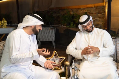 تبادل الاحاديث والاخبار بتعابير السرور، زينة واضاءة رمضانية، الجلسة الليلة الخارجية في احدى شاليهات الامارات، حسن الاستقبال والكرم في شهر رمضان المبارك، صديقان عربيان خليجيان اماراتيان يرتديان الكندورة والغترة يجلسان في الهواء الطلق