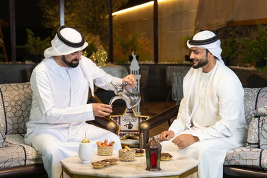 منتج وطني محلي اماراتي، زينة واضاءة رمضانية، عادات وتقاليد شرب القهوة التقليدية، الجلسة الليلة الخارجية في احدى شاليهات الامارات، حسن الاستقبال والكرم في شهر رمضان المبارك، صديقان عربيان خليجيان اماراتيان يرتديان الكندورة والغترة يجلسان في الهواء الطلق
