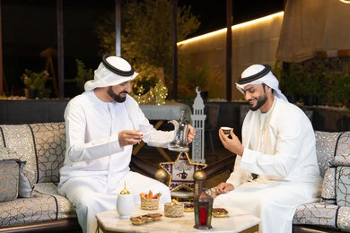 الجلسة الليلة الخارجية في احدى شاليهات الامارات، حسن الاستقبال والكرم في شهر رمضان المبارك، صديقان عربيان خليجيان اماراتيان يرتديان الكندورة والغترة يجلسان في الهواء الطلق، منتج وطني محلي اماراتي، زينة واضاءة رمضانية، عادات وتقاليد شرب القهوة التقليدية