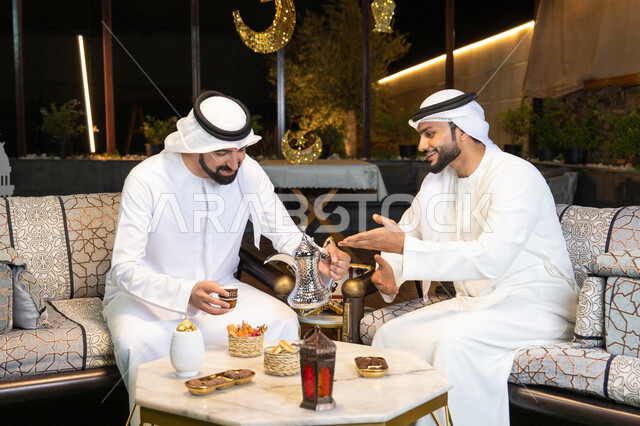 الجلسة الليلة الخارجية في احدى شاليهات الامارات، حسن الاستقبال والكرم في شهر رمضان المبارك، صديقان عربيان خليجيان اماراتيان يرتديان الكندورة والغترة يجلسان في الهواء الطلق، منتج وطني محلي اماراتي، زينة واضاءة رمضانية، عادات وتقاليد شرب القهوة التقليدية