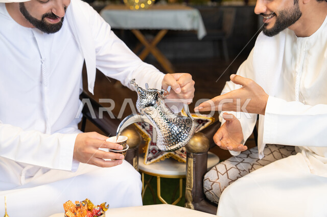 زينة واضاءة رمضانية، عادات وتقاليد شرب القهوة التقليدية، الجلسة الليلة الخارجية في احدى شاليهات الامارات، حسن الاستقبال والكرم في شهر رمضان المبارك، صديقان عربيان خليجيان اماراتيان يرتديان الكندورة والغترة يجلسان في الهواء الطلق، منتج وطني محلي اماراتي