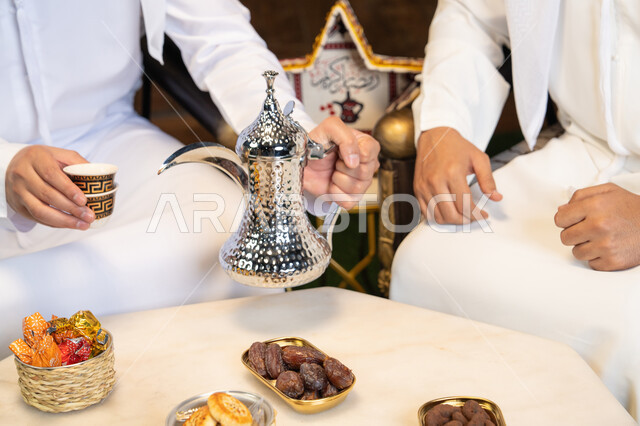 تقاليد وعادات شرب القهوة التقليدية، الجلسة الليلة الخارجية في احدى شاليهات الامارات، حسن الاستقبال والكرم في شهر رمضان المبارك، صديقان عربيان خليجيان اماراتيان يرتديان الكندورة والغترة يجلسان في الهواء الطلق، منتج وطني محلي اماراتي، زينة واضاءة رمضانية