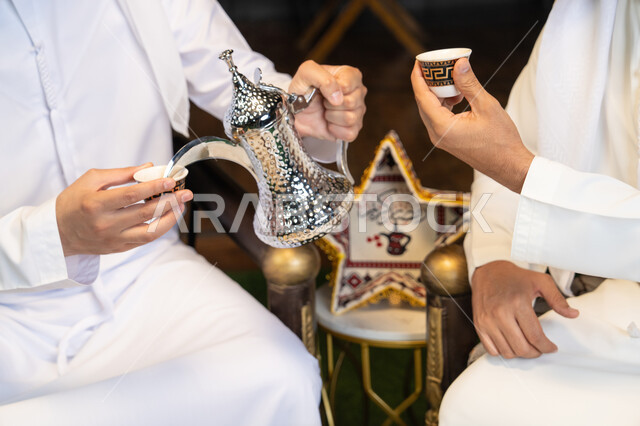 زينة واضاءة رمضانية، عادات وتقاليد شرب القهوة التقليدية، الجلسة الليلة الخارجية في احدى شاليهات الامارات، حسن الاستقبال والكرم في شهر رمضان المبارك، صديقان عربيان خليجيان اماراتيان يرتديان الكندورة والغترة يجلسان في الهواء الطلق، منتج وطني محلي اماراتي