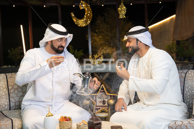 الجلسة الليلة الخارجية في احدى شاليهات الامارات، حسن الاستقبال والكرم في شهر رمضان المبارك، صديقان عربيان خليجيان اماراتيان يرتديان الكندورة والغترة يجلسان في الهواء الطلق، منتج وطني محلي اماراتي، زينة واضاءة رمضانية، عادات وتقاليد شرب القهوة التقليدية