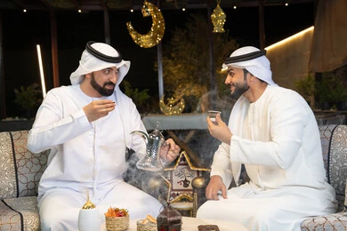 الجلسة الليلة الخارجية في احدى شاليهات الامارات، حسن الاستقبال والكرم في شهر رمضان المبارك، صديقان عربيان خليجيان اماراتيان يرتديان الكندورة والغترة يجلسان في الهواء الطلق، منتج وطني محلي اماراتي، زينة واضاءة رمضانية، عادات وتقاليد شرب القهوة التقليدية