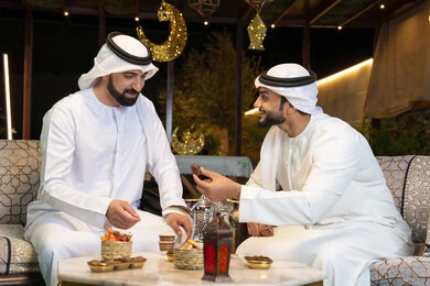تناول التمر السكري الطازج، منتج وطني محلي اماراتي، زينة واضاءة رمضانية، الجلسة الليلة الخارجية في احدى شاليهات الامارات، حسن الاستقبال والكرم في شهر رمضان المبارك، صديقان عربيان خليجيان اماراتيان يرتديان الكندورة والغترة يجلسان في الهواء الطلق