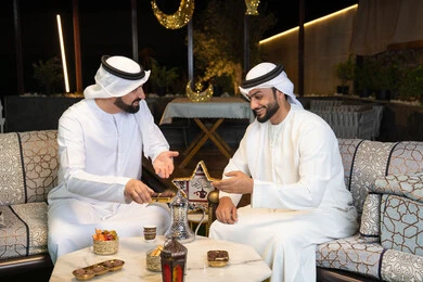 حسن الاستقبال والكرم في شهر رمضان المبارك، صديقان عربيان خليجيان اماراتيان يرتديان الكندورة والغترة يجلسان في الهواء الطلق، تناول التمر السكري الطازج، منتج وطني محلي اماراتي، زينة واضاءة رمضانية، الجلسة الليلة الخارجية في احدى شاليهات الامارات