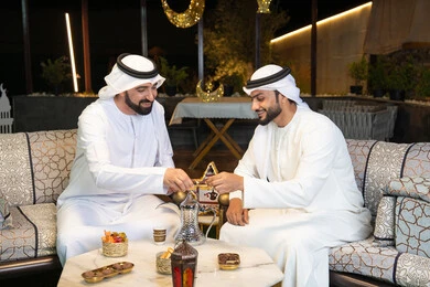 ضيافة  التمر السكري الطازج، منتج وطني محلي اماراتي، زينة واضاءة رمضانية، الجلسة الليلة الخارجية في احدى شاليهات الامارات، حسن الاستقبال والكرم في شهر رمضان المبارك، صديقان عربيان خليجيان اماراتيان يرتديان الكندورة والغترة يجلسان في الهواء الطلق