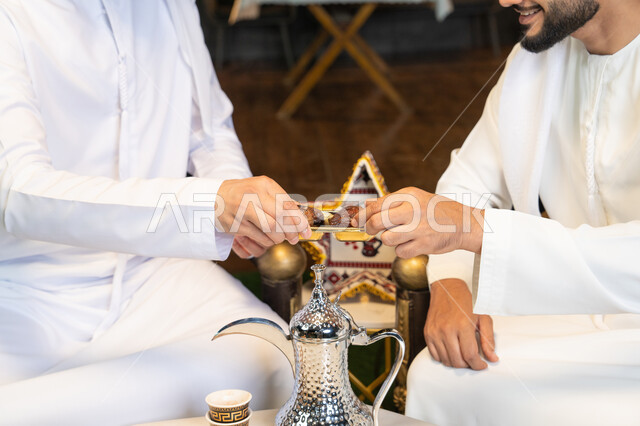 ضيافة  التمر السكري الطازج، منتج وطني محلي اماراتي، زينة واضاءة رمضانية، الجلسة الليلة الخارجية في احدى شاليهات الامارات، حسن الاستقبال والكرم في شهر رمضان المبارك، صديقان عربيان خليجيان اماراتيان يرتديان الكندورة والغترة يجلسان في الهواء الطلق