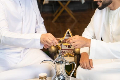 ضيافة  التمر السكري الطازج، منتج وطني محلي اماراتي، زينة واضاءة رمضانية، الجلسة الليلة الخارجية في احدى شاليهات الامارات، حسن الاستقبال والكرم في شهر رمضان المبارك، صديقان عربيان خليجيان اماراتيان يرتديان الكندورة والغترة يجلسان في الهواء الطلق