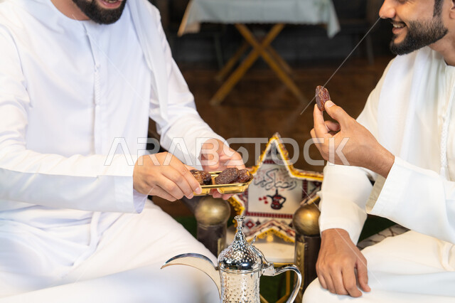 تناول التمر السكري الطازج، منتج وطني محلي اماراتي، زينة واضاءة رمضانية، الجلسة الليلة الخارجية في احدى شاليهات الامارات، حسن الاستقبال والكرم في شهر رمضان المبارك، صديقان عربيان خليجيان اماراتيان يرتديان الكندورة والغترة يجلسان في الهواء الطلق