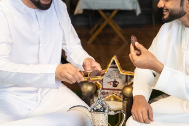تناول التمر السكري الطازج، منتج وطني محلي اماراتي، زينة واضاءة رمضانية، الجلسة الليلة الخارجية في احدى شاليهات الامارات، حسن الاستقبال والكرم في شهر رمضان المبارك، صديقان عربيان خليجيان اماراتيان يرتديان الكندورة والغترة يجلسان في الهواء الطلق