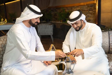 حسن الاستقبال والكرم في شهر رمضان المبارك، صديقان عربيان خليجيان اماراتيان يرتديان الكندورة والغترة يجلسان في الهواء الطلق، تناول التمر السكري الطازج، منتج وطني محلي اماراتي، زينة واضاءة رمضانية، الجلسة الليلة الخارجية في احدى شاليهات الامارات
