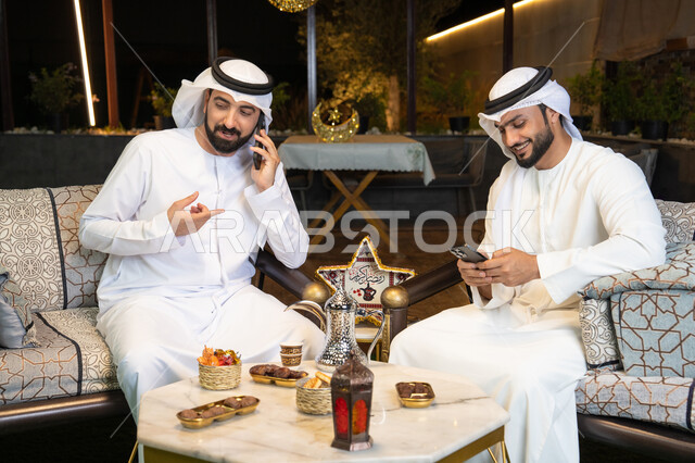 An outdoor evening session in one of the chalets in the Emirates, warm welcome and hospitality in the holy month of Ramadan, two Emirati Gulf Arab friends wearing kandura and ghutra sitting in the open air, making a phone call using a mobile phone with expressions of pleasure, Ramadan decorations and lighting