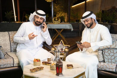 An outdoor evening session in one of the chalets in the Emirates, warm welcome and hospitality in the holy month of Ramadan, two Emirati Gulf Arab friends wearing kandura and ghutra sitting in the open air, making a phone call using a mobile phone with expressions of pleasure, Ramadan decorations and lighting