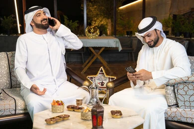 اجراء مكالمة هاتفية باستخدام الهاتف المحمول بتعابير السرور، زينة واضاءة رمضانية، الجلسة الليلة الخارجية في احدى شاليهات الامارات، حسن الاستقبال وكرم الضيافة في شهر رمضان المبارك، صديقان عربيان خليجيان اماراتيان يرتديان الكندورة والغترة يجلسان في الهواء الطلق