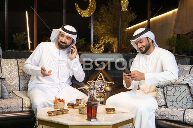 الجلسة الليلة الخارجية في احدى شاليهات الامارات، حسن الاستقبال وكرم الضيافة في شهر رمضان المبارك، صديقان عربيان خليجيان اماراتيان يرتديان الكندورة والغترة يجلسان في الهواء الطلق ، اجراء مكالمة هاتفية باستخدام الهاتف المحمول بتعابير السرور، زينة واضاءة رمضانية