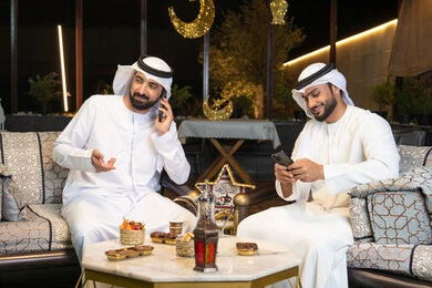 الجلسة الليلة الخارجية في احدى شاليهات الامارات، حسن الاستقبال وكرم الضيافة في شهر رمضان المبارك، صديقان عربيان خليجيان اماراتيان يرتديان الكندورة والغترة يجلسان في الهواء الطلق ، اجراء مكالمة هاتفية باستخدام الهاتف المحمول بتعابير السرور، زينة واضاءة رمضانية