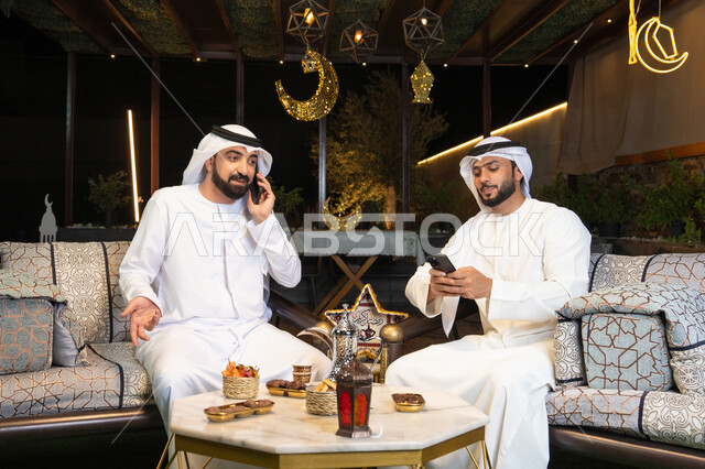اجراء مكالمة هاتفية باستخدام الهاتف المحمول بتعابير السرور، زينة واضاءة رمضانية، الجلسة الليلة الخارجية في احدى شاليهات الامارات، حسن الاستقبال وكرم الضيافة في شهر رمضان المبارك، صديقان عربيان خليجيان اماراتيان يرتديان الكندورة والغترة يجلسان في الهواء الطلق