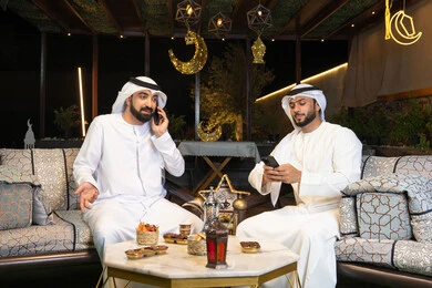 اجراء مكالمة هاتفية باستخدام الهاتف المحمول بتعابير السرور، زينة واضاءة رمضانية، الجلسة الليلة الخارجية في احدى شاليهات الامارات، حسن الاستقبال وكرم الضيافة في شهر رمضان المبارك، صديقان عربيان خليجيان اماراتيان يرتديان الكندورة والغترة يجلسان في الهواء الطلق